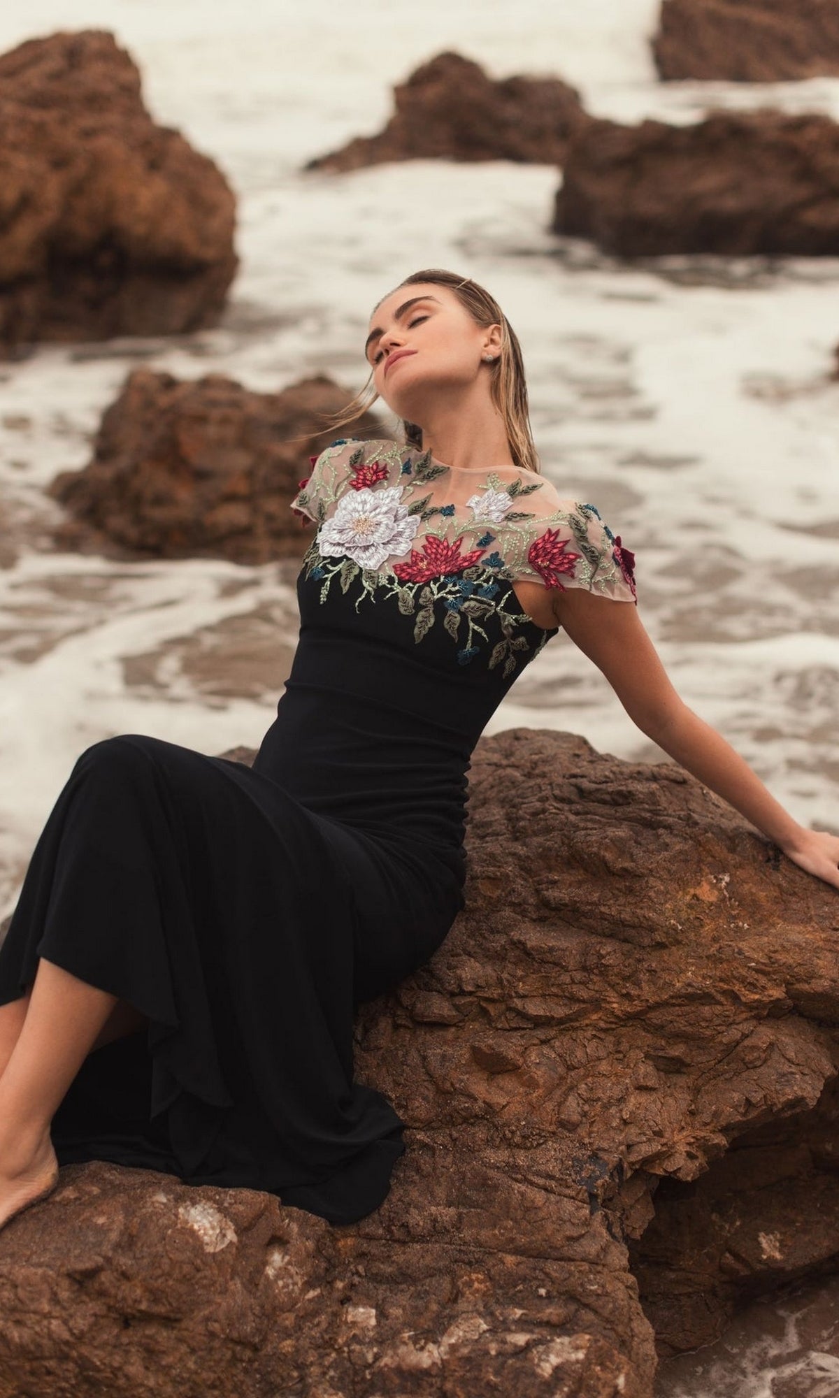 A woman wearing the Mac Duggal 11817 floral long black formal dress sits barefoot on a rock by the ocean, with waves and rocks in the background.